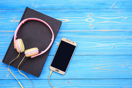 Top view smartphone, new modern pink music headphone and book on blue wooden plank background. With space for text or designの写真素材
