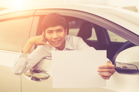 Young asian businessman sitting in the modern car and holding blank of white paper or business card. For car rent or car business. Vintage color and warm lens flare filter effectの写真素材