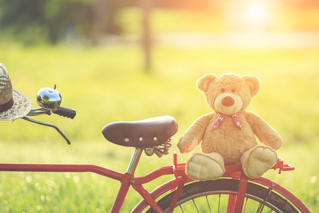 Close up lovely brown teddy bear in rattan basket on vintage bike in green field with lens flare. Warm toning effect. Retro and vintage styleの写真素材