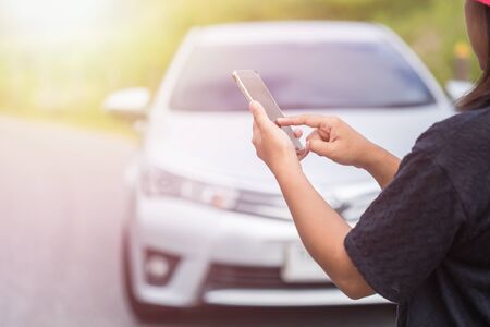 Asian woman using smartphone in front of her broken car on the road. Contacting car technician or...