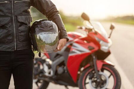 Handsome motorcyclist wear leather jacket and holding helmet on the road. Safe ride and transportation concept