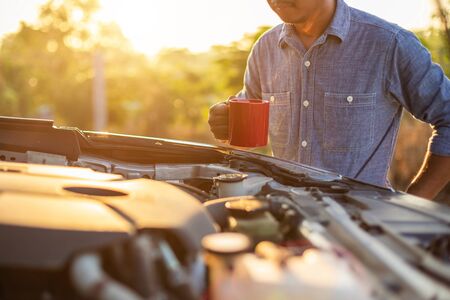 Asian man holding a red coffee cup and checking the engine of his car in the morning. Car maintenance or service before driving conceptの写真素材