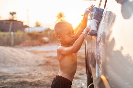 Happy asian little boy playing white soap and using blue sponge to washing the car at outdoor in sunset time. Kid and family activity conceptの写真素材