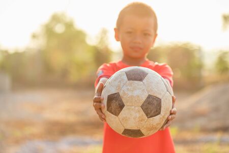 Young Asian boy playing old and dirty classic soccer ball at the countryside in the morning with sunlight effectの写真素材