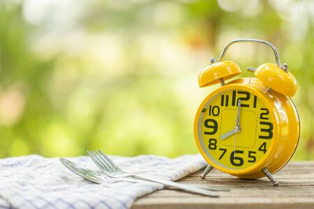 Close up yellow alarm clock, Fork, and spoon on wooden table with green outdoor nature blur...