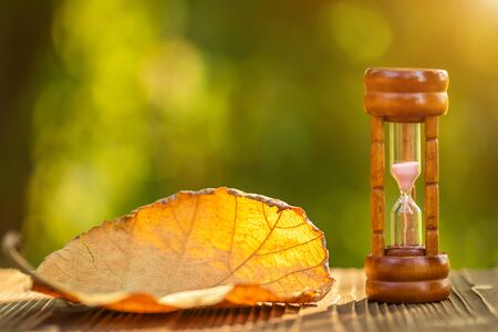 Close up vintage hourglass or sandglass with dry tree leaf on wooden table, Green nature blur backgroundの写真素材