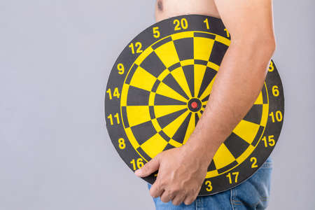 Fat people holding round yellow dartboard beside his belly position. Target of losing weight concept. Studio shot isolated on grey backgroundの写真素材