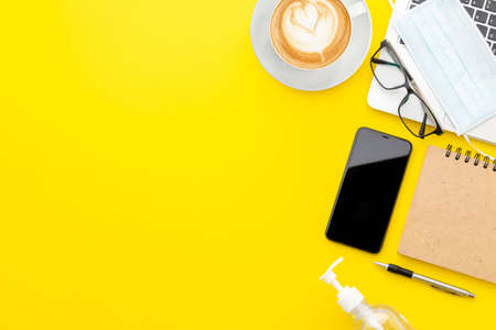Top view new preventive mask, laptop, eyeglasses, smartphone, hand sanitizer gel, coffee cup and book on yellow background with copy space for text or design. Coronavirus disease (COVID-19) or Healthy conceptの写真素材