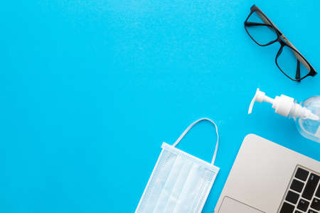 Top view new preventive mask, sanitizer gel, laptop and eye glasses on blue background with copy space for text or design. Coronavirus disease (COVID-19) or Healthy conceptの写真素材