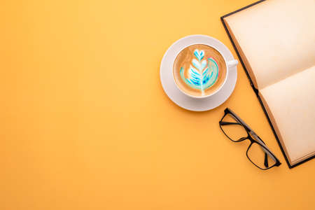 Top view white cup of hot Latte art coffee, book and eyeglasses on yellow background. Copy space for...