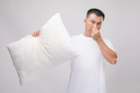 The pillow smells bad. Portrait Asian man holding white pillow and getting bad smell. Studio shot on grey backgroundの写真素材