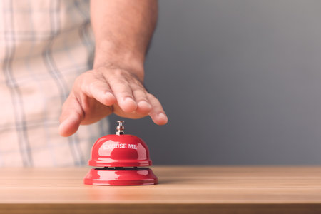 Close up hand ringing the red call bell or service bell ring on the brown wooden desk with grey wall background. For hotel or restaurant advertising conceptの写真素材