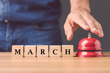 Close up hand ringing the red call bell and wooden blocks write word MARCH arranged on wooden desk...