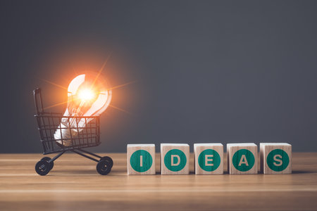 IDEAS write on wooden block arranged on wooden table and light bulb in shopping trolley on grey wall background. Start up or Create the new idea concept.の写真素材