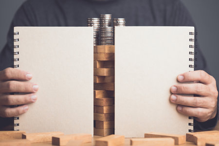 Hidden about risks in business or financial. Idea to keep risky as secret. Businessman using book to hide tower wooden block stacking game on wooden desk in office. Studio shot.の写真素材