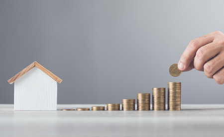 Man is stacking coins on a table, arranging them from smallest to largest, with a wooden house model beside him. Emphasizes the concept of saving money for future investments and financial stabilityの写真素材