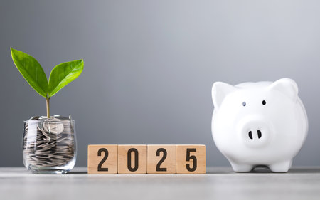 2025 written on a wooden block next to a glass jar filled with coins and white piggy bank placed on...