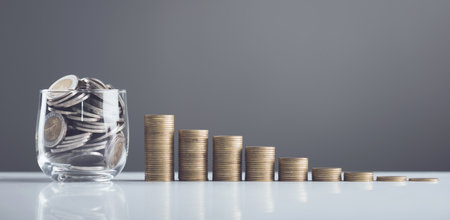 Coins in a glass jar, with stacks of coins arranged from smallest to largest on the table. The grey background highlights the concepts of saving, financial planning, and steady financial growthの写真素材