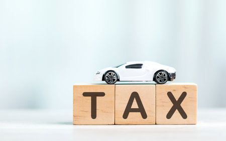 TAX is written on a wooden block next to a car model and a jar filled with coins and a small plant on a wooden table. Car tax awareness, financial planning, savings, and responsible money managementの写真素材