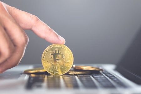 Man holds a golden Bitcoin while using a laptop, representing Bitcoin as a form of digital currency...