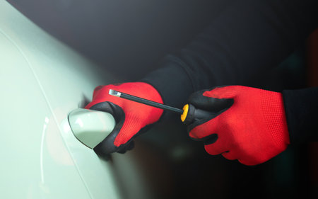 A man's gloved hand holding a screwdriver, attempting to pry open a car door handle. The image...