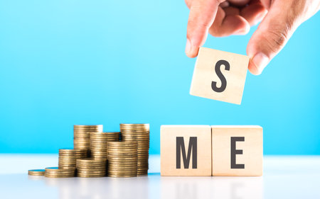 A hand stacking coins with 'SME' written on a wooden block and more coins on the table. Represents financial growth of small and medium-sized enterprises, emphasizing investment and money managementの写真素材