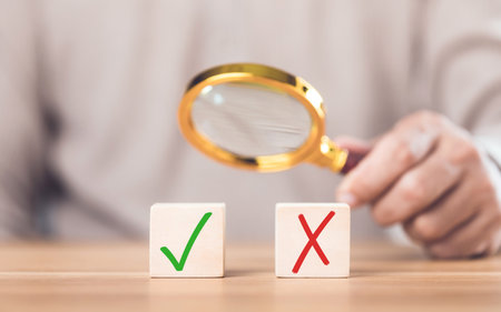 Man holding a magnifying glass over checkmark and 'X' symbols on wooden blocks. The image reflects the concept of choosing right from wrong, highlighting careful decision-making and evaluationの写真素材