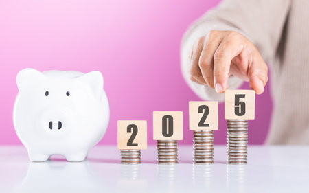 A businessperson arranges wooden blocks labeled '2025' on a rising stack of coins next to a white piggy bank, symbolizing financial growth, planning, and saving for future successの写真素材