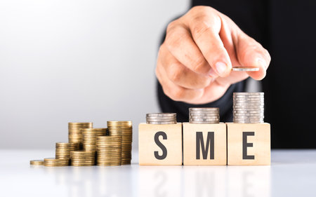 A hand stacking coins with 'SME' written on a wooden block and more coins on the table. Represents financial growth of small and medium-sized enterprises, emphasizing investment and money managementの写真素材