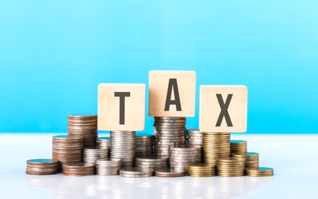 TAX is write on a wooden block next to a stack of coins. Against light blue background. Tax awareness, financial planning, savings, and effective money managementの写真素材