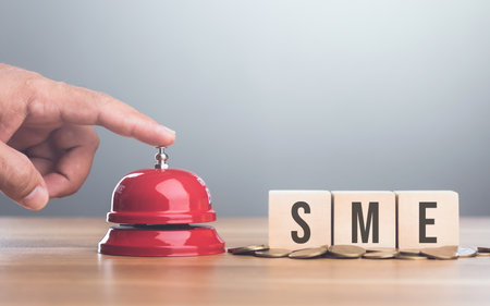 Hand presses a red bell next to a stack of coins and a wooden block labeled 'SME,' symbolizing the start of a new small business venture and the importance of early financial planningの写真素材