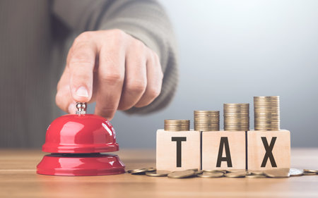 A hand presses a red bell beside a stack of coins on a wooden block labeled TAX, An urgent call to address tax matters and financial planning, Timely tax payments and financial responsibilityの写真素材