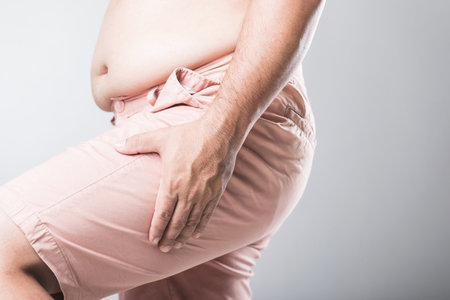A man grips his thigh with both hands, showing discomfort from muscle pain, possibly related to excess weight. Men's health, particularly the impact of obesity on muscle strain and overall well-beingの写真素材