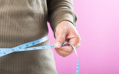 A man measuring his waistline with a tape measure, emphasizing the importance of regular health checks and awareness of weight management to promote better overall healthの写真素材