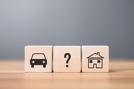 A man contemplates wooden blocks labeled with car and house symbols, representing the decision between purchasing a car or a home first. Reflects thoughtful financial planning and prioritizationの写真素材