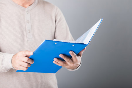 A man holds a blue life insurance policy booklet, reading and reviewing its details against a gray background. This image highlights the importance of understanding and planning financial securityの写真素材