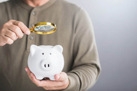 Man holds a white piggy bank and uses a magnifying glass to examine it, symbolizing the careful study of savings, financial planning, and investment strategies for future growthの写真素材