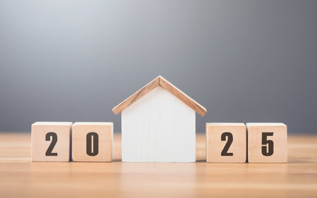 2025 is write on a wooden block next to glass jar filled with coins and a house model, placed on a wooden table against a bright white. Financial planning, savings, and effective money managementの写真素材