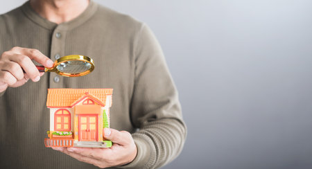 Homeowner holds a house model and uses a magnifying glass to examine it, symbolizing the process of finding the right home price. Reflects the careful consideration in buying or selling real estateの写真素材