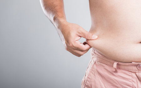 A man with a larger physique stands, holding his waist to check or measure his waistline and abdominal fat, emphasizing the importance of monitoring body shape and men's healthの写真素材