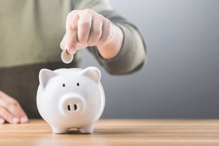 A man drops a coin into a white piggy bank on a table, symbolizing the practice of saving money. This image reflects financial responsibility and future planningの写真素材