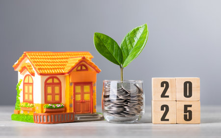 A coin jar with a sprouting plant symbolizes financial growth, alongside wooden blocks reading '2025' and a house model. This image represents saving for real estate goals in the coming yearの写真素材