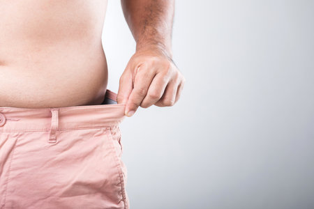 A man with a larger physique stands adjusting the waistband of his pants to check if they are tight or loose, reflecting the importance of monitoring body shape and overall health for menの写真素材