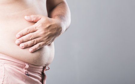 A man presses his hand on his left abdomen, showing discomfort potentially linked to kidney issues caused by excess weight. Image emphasizes men's health and the impact of obesity on internal organsの写真素材