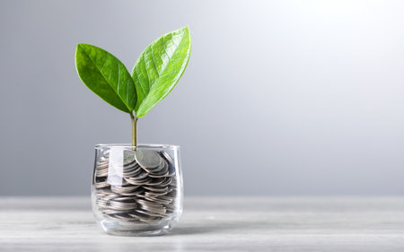 Glass jar filled with coins, with a small seedling growing in the center, placed on a table against a dark grey backdrop. The image represents the concept of saving and financial growth for futureの写真素材