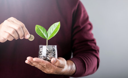 A businessman holds a coin jar with a sprouting plant, shielding it with his other hand. The gesture symbolizes nurturing and protecting personal finances for sustainable growthの写真素材
