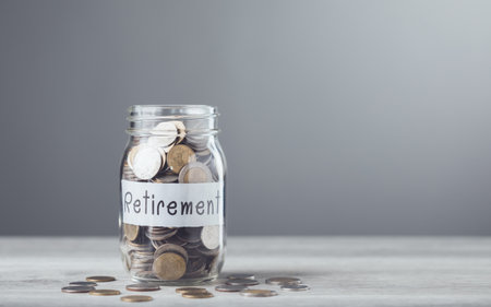 A glass jar filled with coins and a sprout labeled 'Retirement,' placed on a table with a gray background, symbolizing saving for financial growth and secure future planningの写真素材