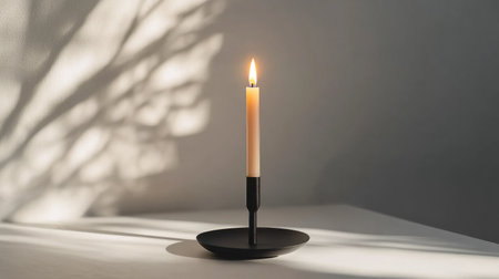 A minimalist black candlestick holding a lit candle, casting a soft glow and shadow on a white surface.の素材