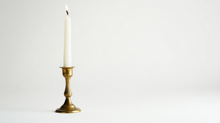 A vintage brass candlestick with a tall white candle, its wax melting gently, on a pristine white background.の素材