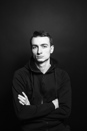 Black and white studio portrait of a young man in a black sweatshirt, arms crossed, seriously looking at the camera, against plain studio backgroundの写真素材